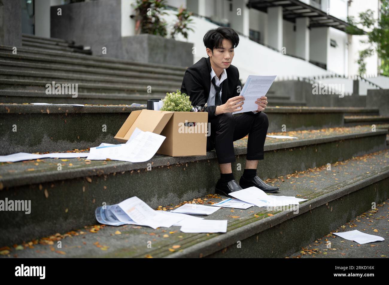 Un giovane uomo d'affari asiatico, stressato e depresso, sta leggendo la sua lettera di licenziamento mentre si siede sulle scale davanti all'ufficio aziendale con un Foto Stock