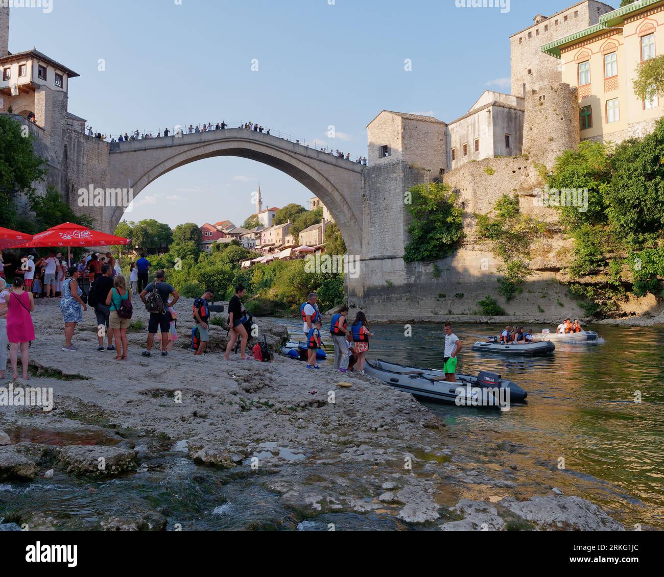 Le persone fanno la fila per un giro in motoscafo su una spiaggia sul fiume Neretva con Stari Most (Ponte Vecchio) alle spalle a Mostar, Bosnia ed Erzegovina, 20 agosto 2023. Foto Stock