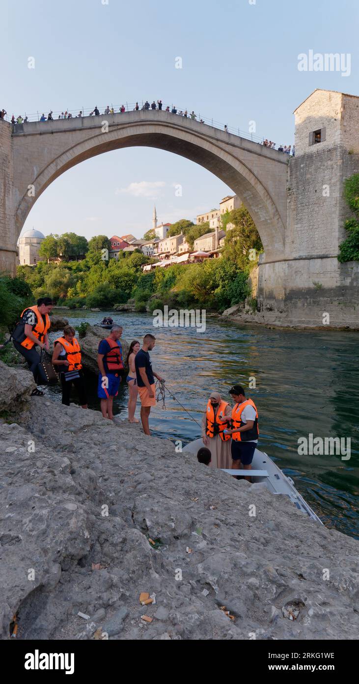 Le persone fanno la fila per un giro in motoscafo su una spiaggia sul fiume Neretva con Stari Most (Ponte Vecchio) alle spalle a Mostar, Bosnia ed Erzegovina, 20 agosto 2023. Foto Stock