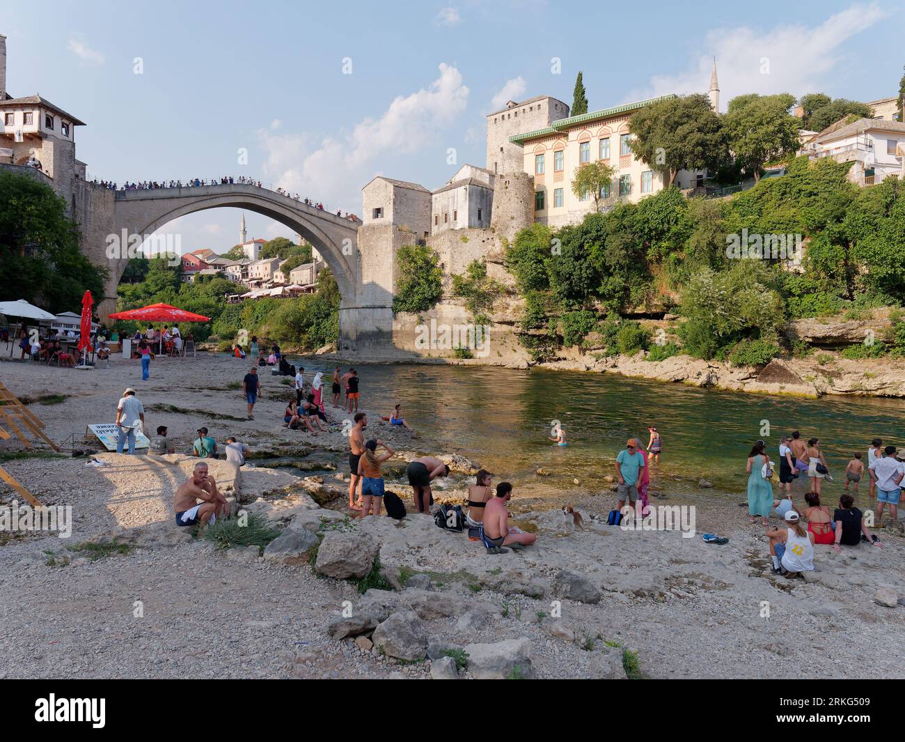 Persone su una spiaggia lungo il fiume Neretva con Stari Most (Ponte Vecchio) alle spalle nella città di Mostar, Bosnia ed Erzegovina, 21 agosto 2023. Foto Stock