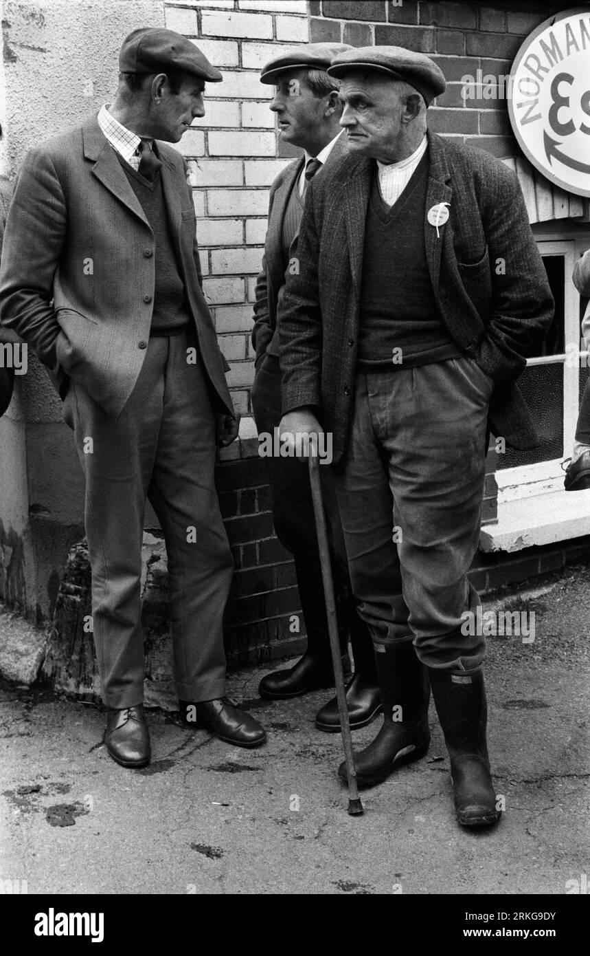 Agricoltori che discutono di un po' di affari nella città di mercato di Hatherleigh Devon 1970s nel Regno Unito. La vendita annuale di cavalli è appena finita, due uomini indossano abiti in tweet, un colletto e cravatta, saltatori (novembre) e scarpe in pelle lucida. Il terzo uomo con bastone da passeggio indossa una vecchia camicia senza colletto, un maglione e una vecchia giacca. Pantaloni a cordoncino e stivali wellington. Tutti e tre gli uomini indossano berretti piatti in tweed. Inghilterra 1973. HOMER SYKES Foto Stock