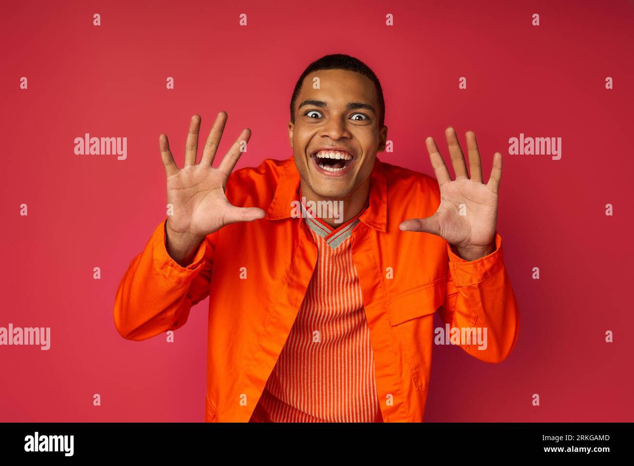 giocoso uomo afroamericano con un'espressione pazzesca che mostra un gesto spaventoso su una camicia rossa e arancione Foto Stock