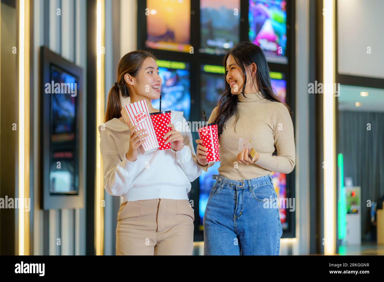 Amici asiatici, popcorn in mano, passeggiate gioiosamente di fronte al cinema, incarnando l'amicizia e l'attesa piena di divertimento per un divertente serata cinematografica Foto Stock