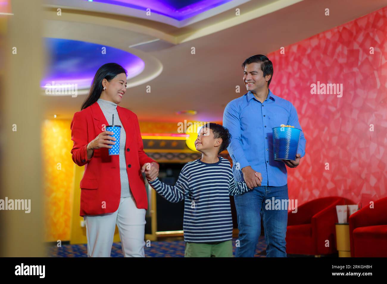 Una famiglia allegra, genitori e figlio in mano, entra nel cinema con bevande e popcorn, pronti a vivere insieme un'esperienza cinematografica commovente. Foto Stock