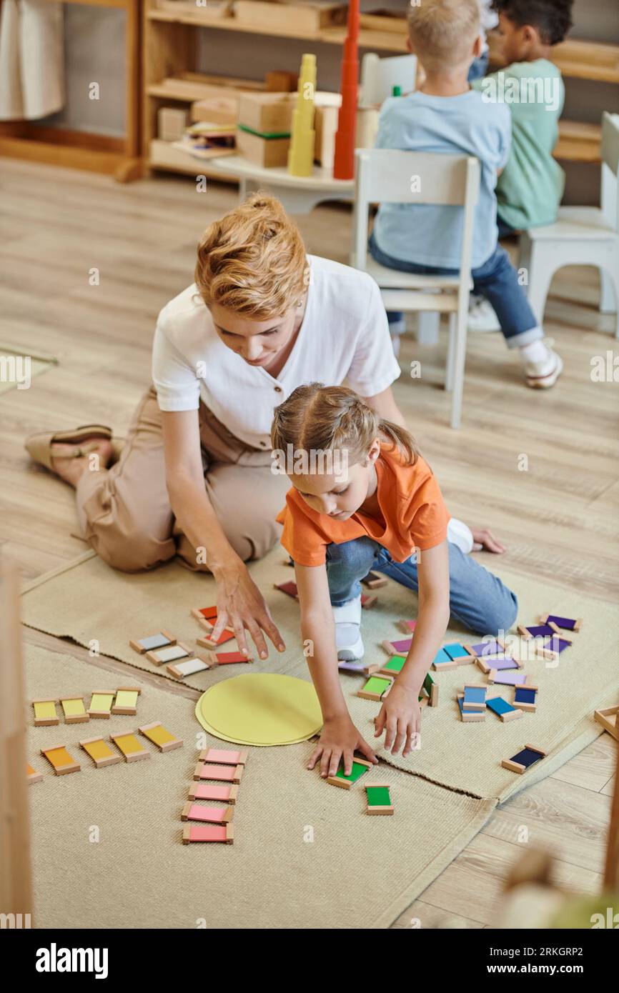 materiale montessori, ragazza che gioca a un gioco di corrispondenza dei colori vicino a un'insegnante gioiosa, ragazzi diversi Foto Stock