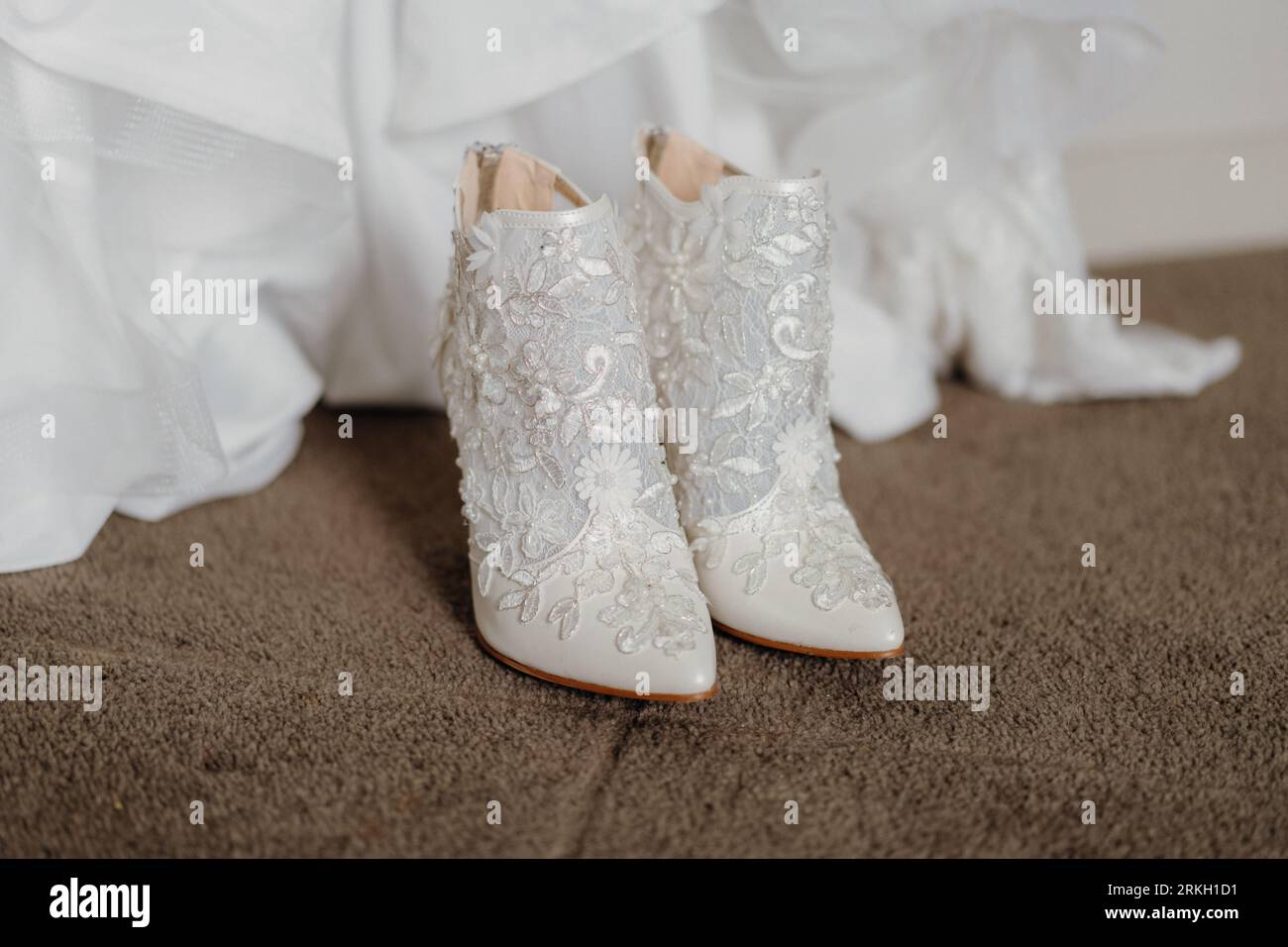 Un paio di scarpe da sposa bianche immacolate su un pavimento in moquette marrone con un abito da sposa sullo sfondo Foto Stock