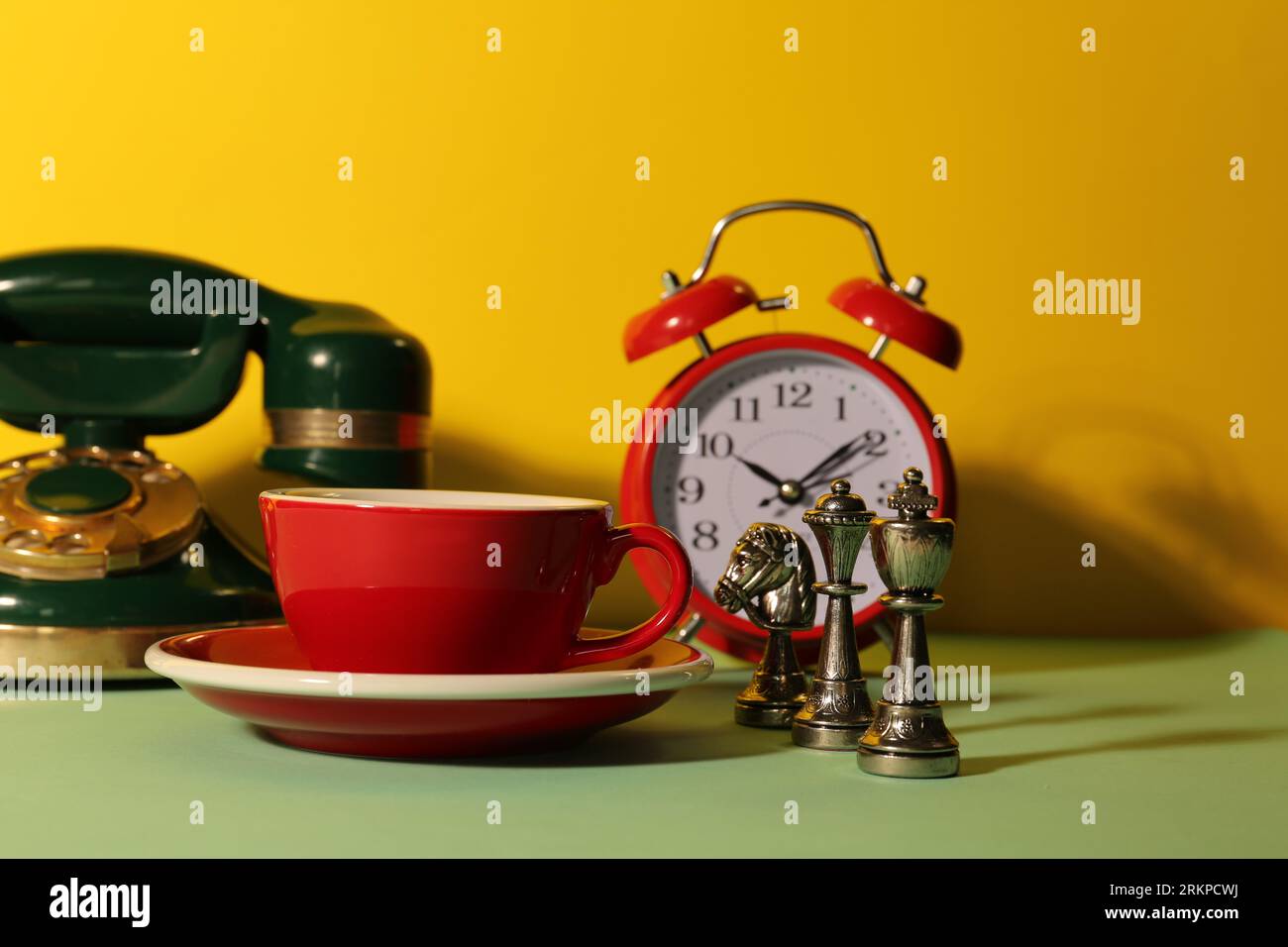 Tazza rossa con piattino, sveglia, telefono vintage e scacchi su un tavolo verde chiaro su sfondo giallo Foto Stock