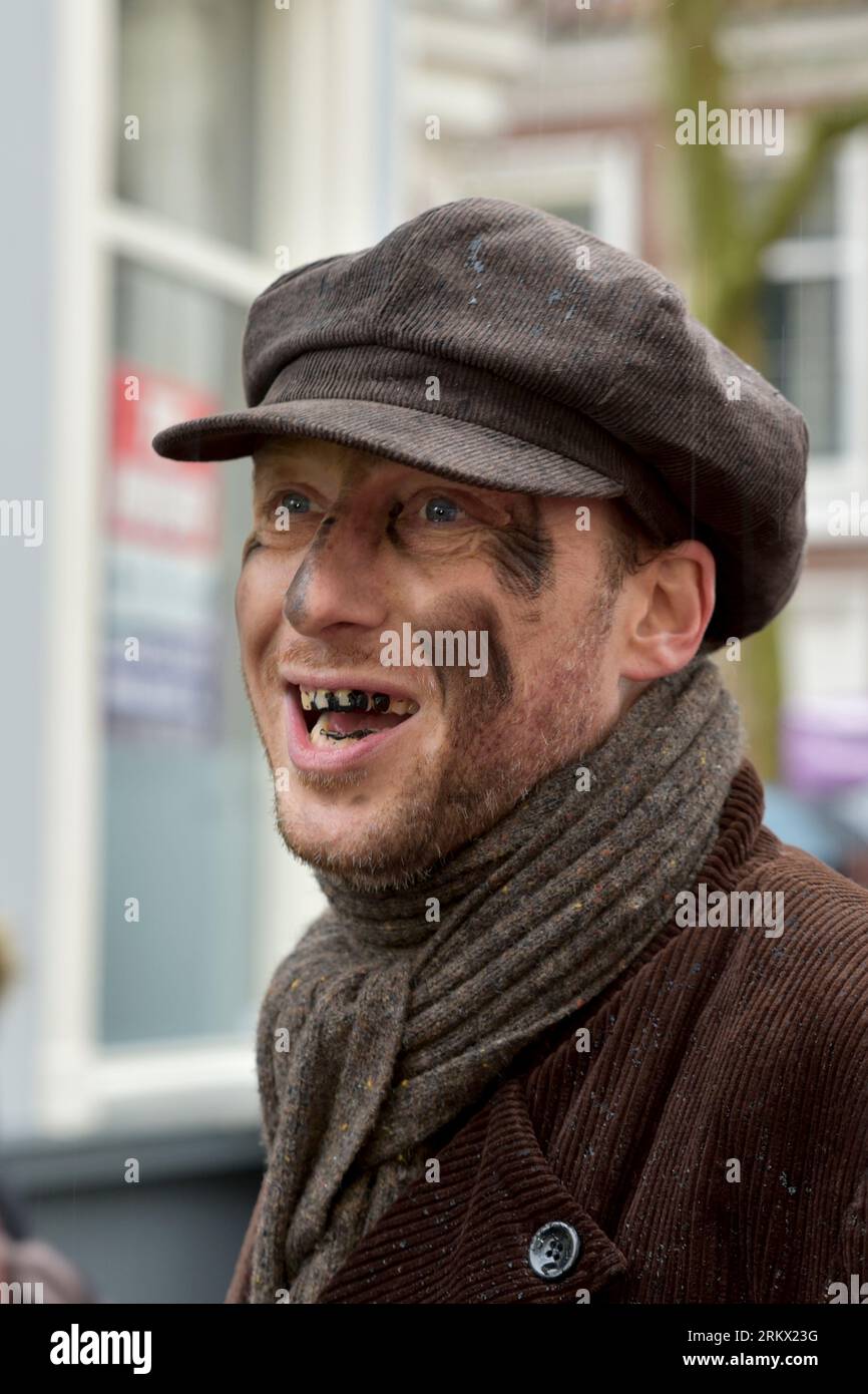 Personaggio con berretto, scialle marrone e giacca, con macchie di fuliggine sul viso e orecchie nere sembra allegramente sorridente durante la Dickens Feast in Deventer Foto Stock