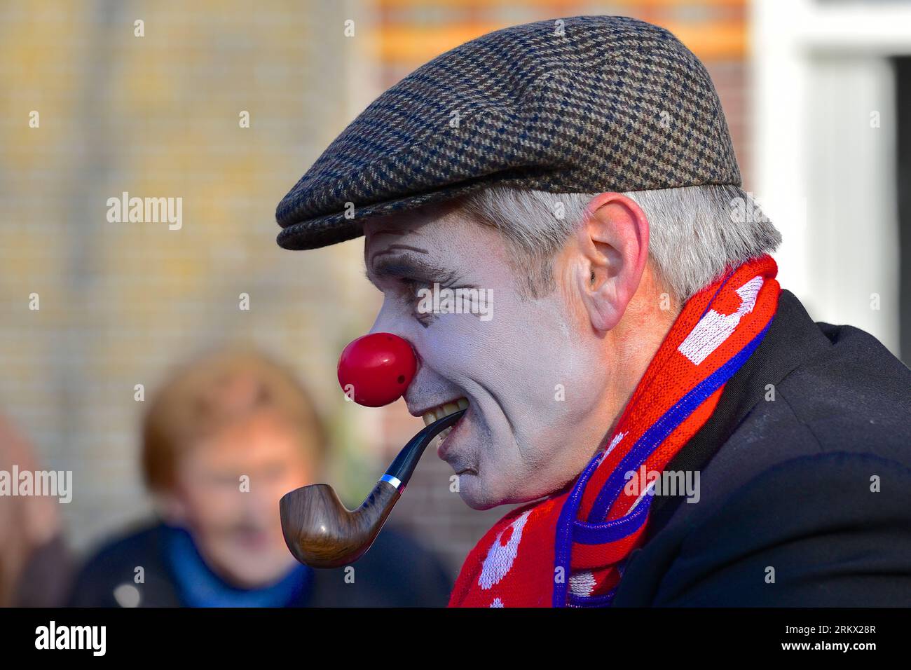 Ritratto di un uomo con un volto dipinto in bianco pallido, naso di un clown, tubo in bocca con un berretto e una sciarpa rosso-blu intorno al collo: carnevale Foto Stock