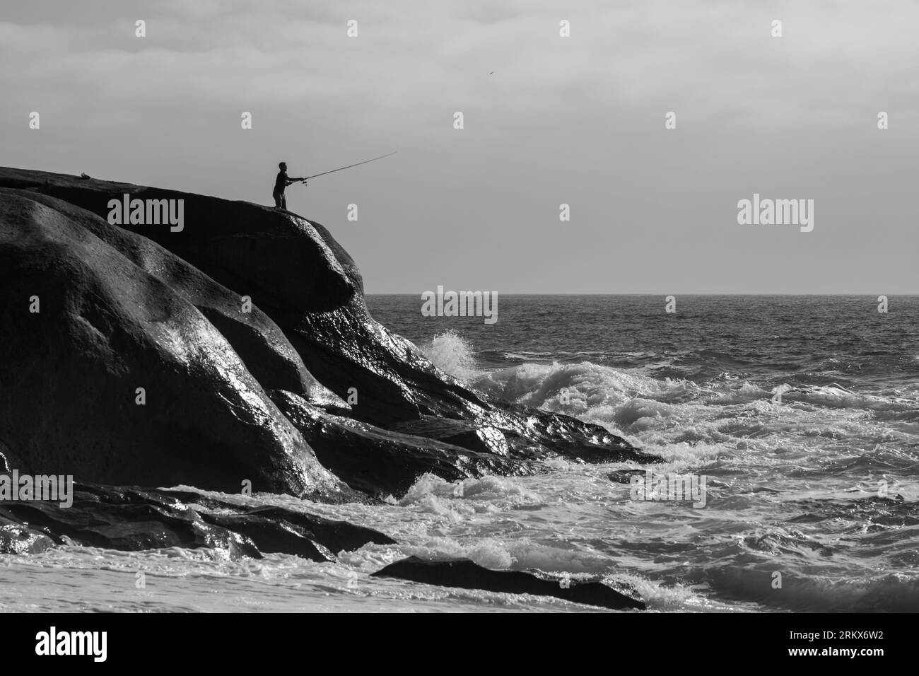 Bianco e nero di una persona in piedi su una costa rocciosa, che pesca nelle acque calme. Foto Stock