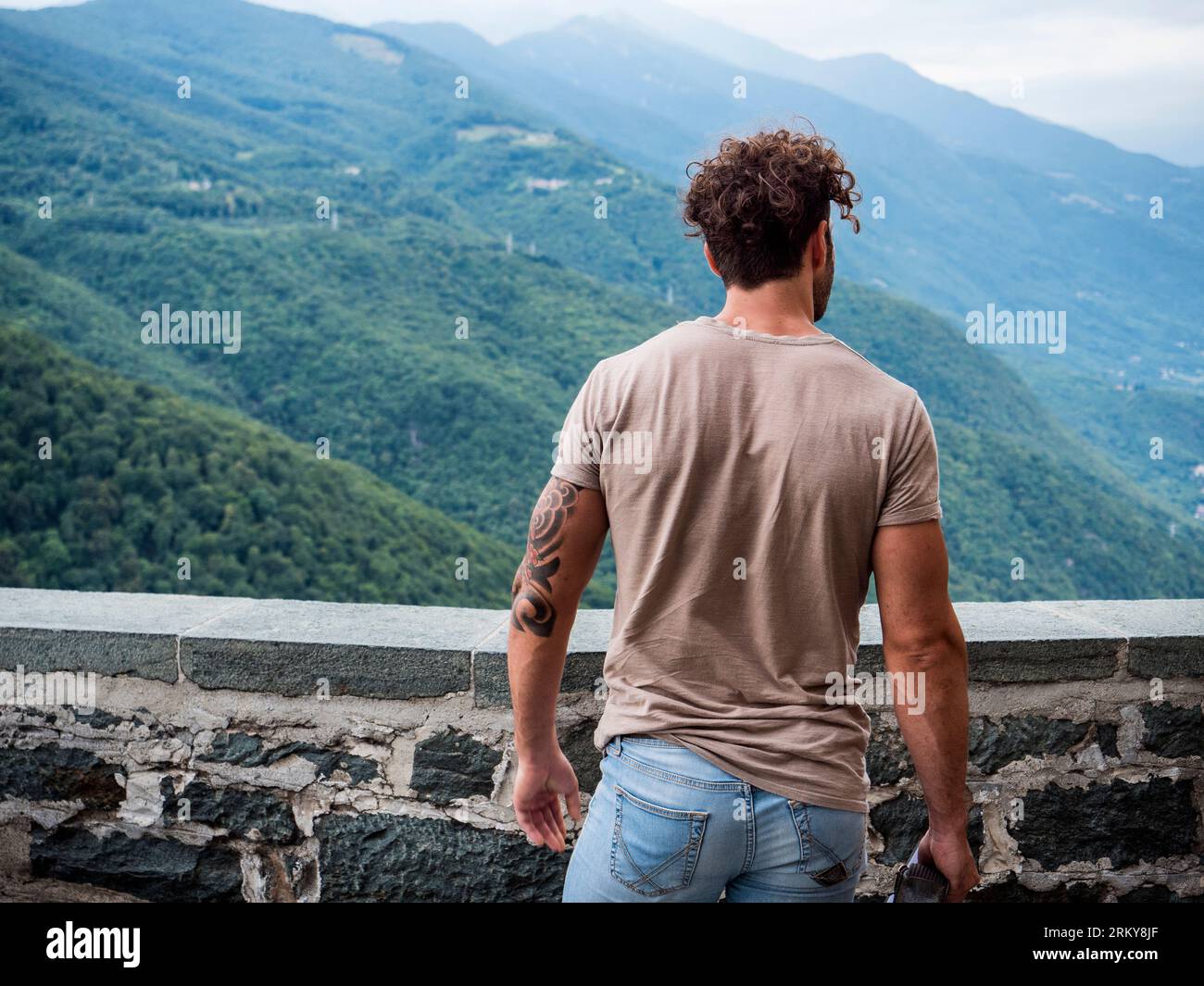 Uomo visto dal retro, in piedi accanto a un muro di pietra con uno sfondo panoramico, che guarda lontano dalla vista. Foto Stock