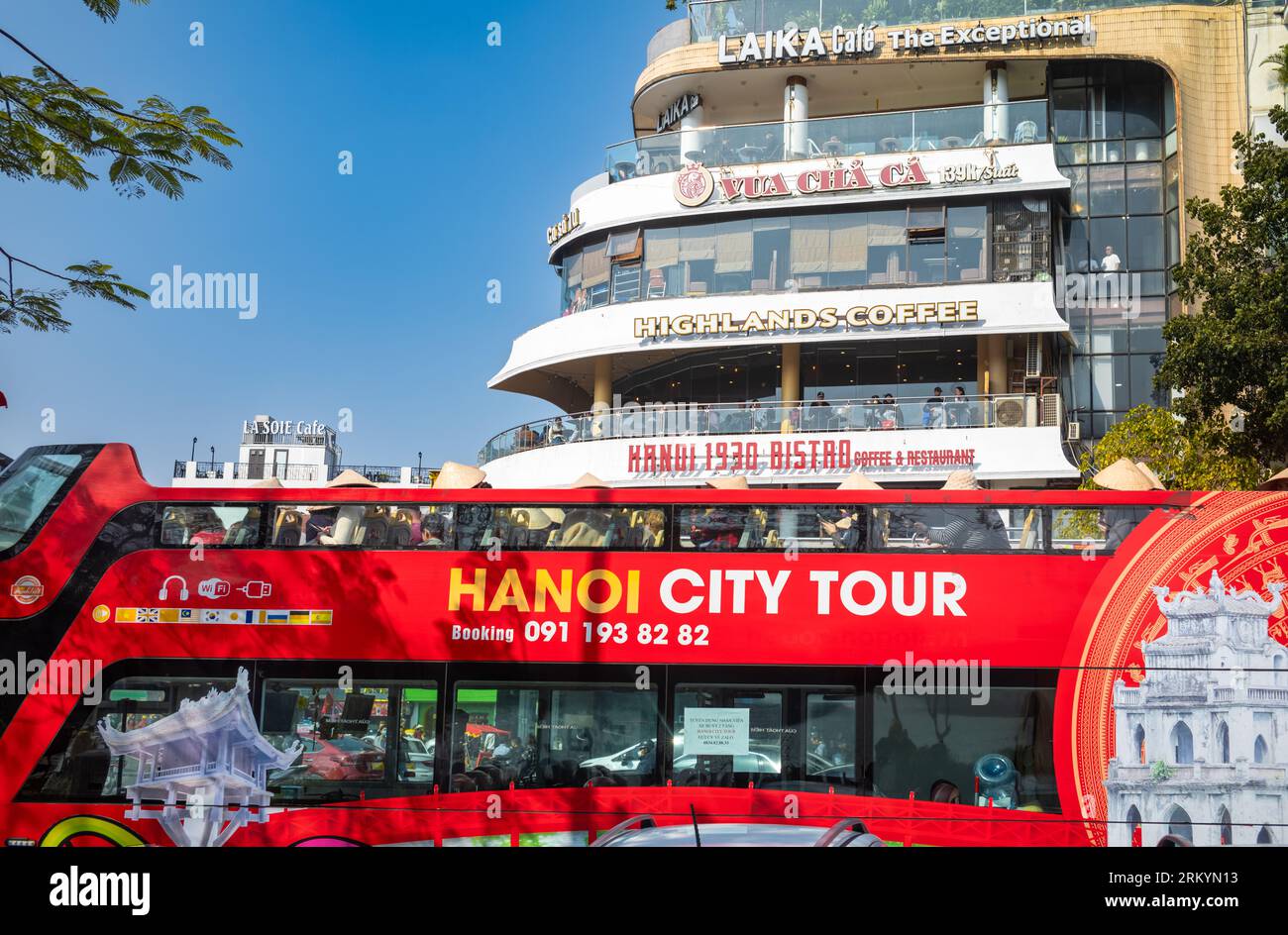 Un autobus rosso a due piani scoperto che offre ai turisti un tour della città di Hanoi passa davanti a diversi famosi caffè alla rotatoria alla fine di Dinh Tien Hoa Foto Stock