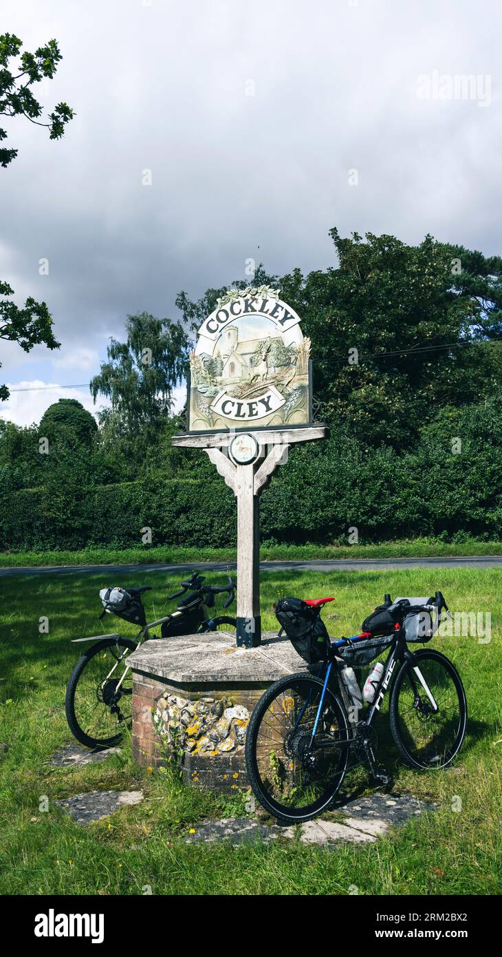 Due biciclette da bikepacking sono parcheggiate di fronte a un cartello del villaggio sul verde a Cockley Cley a Norfolk, Inghilterra, Regno Unito Foto Stock