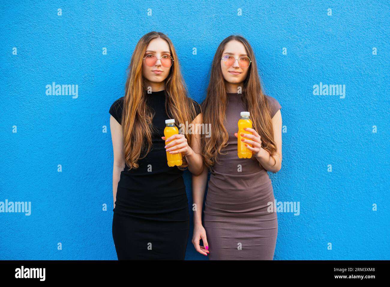 Giovani sorelle gemelle con fantastici occhiali da sole che contengono bottiglie di plastica limonata gialle su sfondo blu Foto Stock