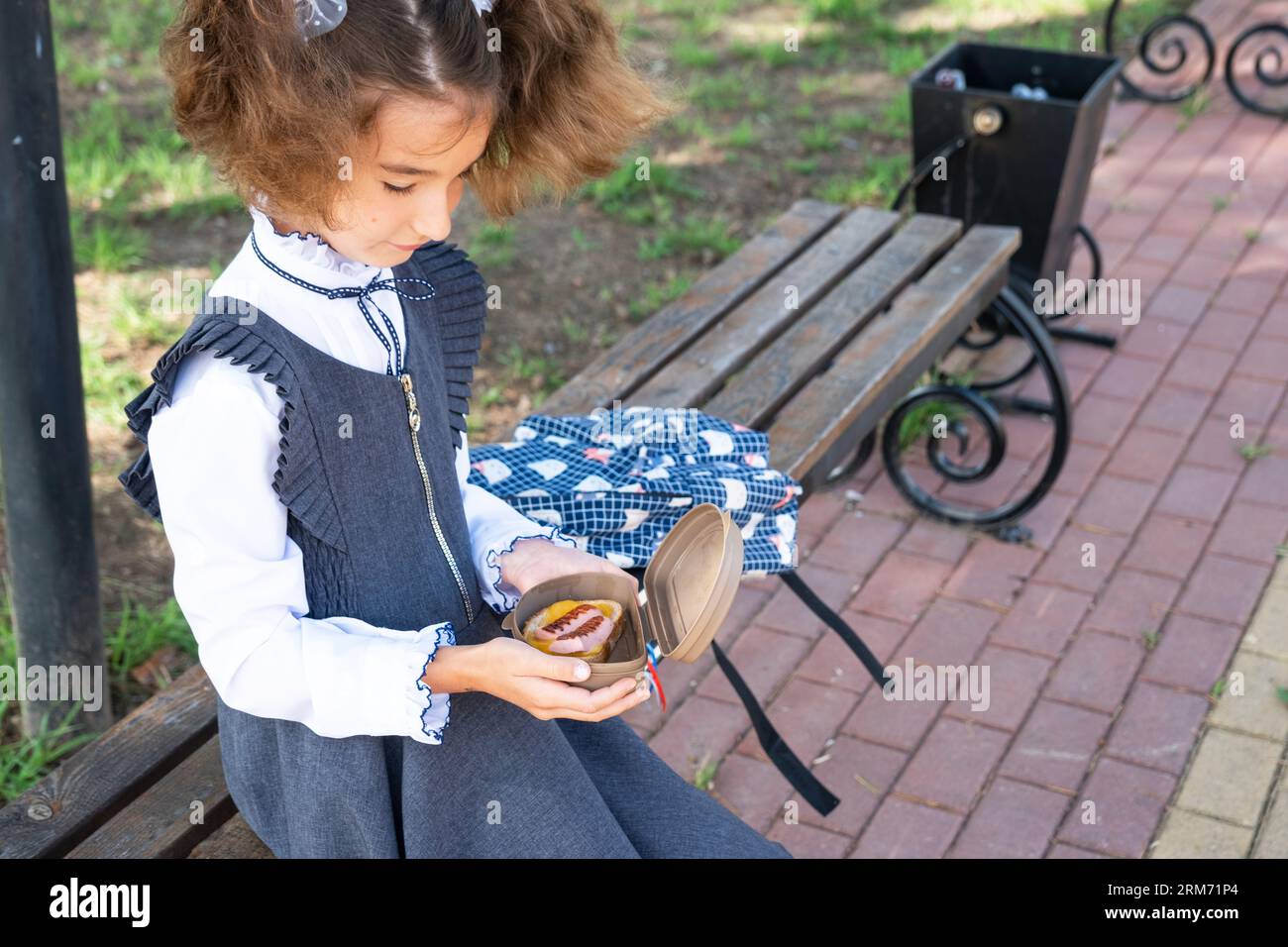 Ragazza con zaino che mangia panino confezionata in una scatola vicino a scuola. Uno spuntino veloce con un panino, cibo malsano, pranzo da scuola. Torna a scuola. Foto Stock