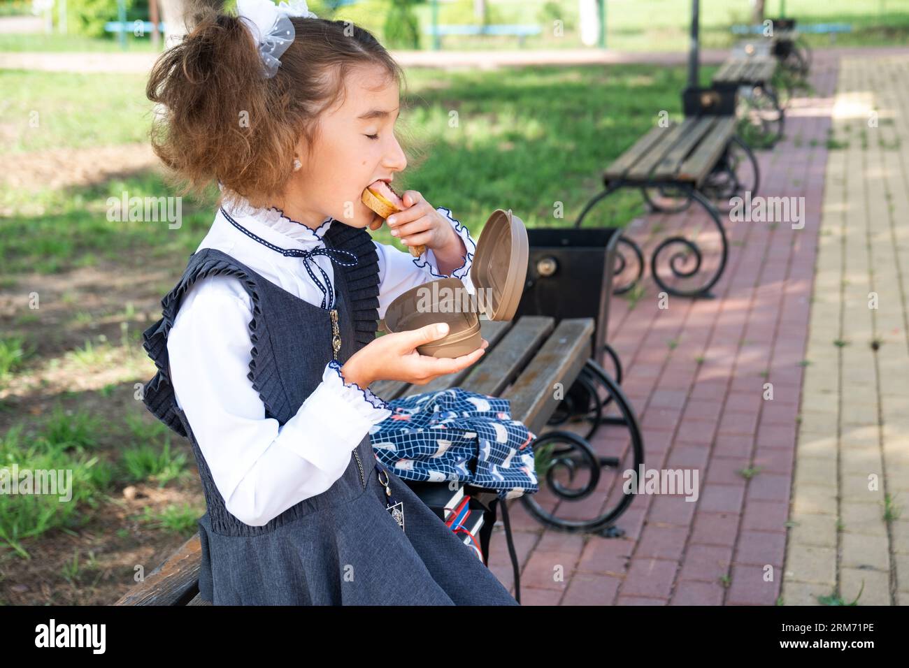 Ragazza con zaino che mangia panino confezionata in una scatola vicino a scuola. Uno spuntino veloce con un panino, cibo malsano, pranzo da scuola. Torna a scuola. Foto Stock