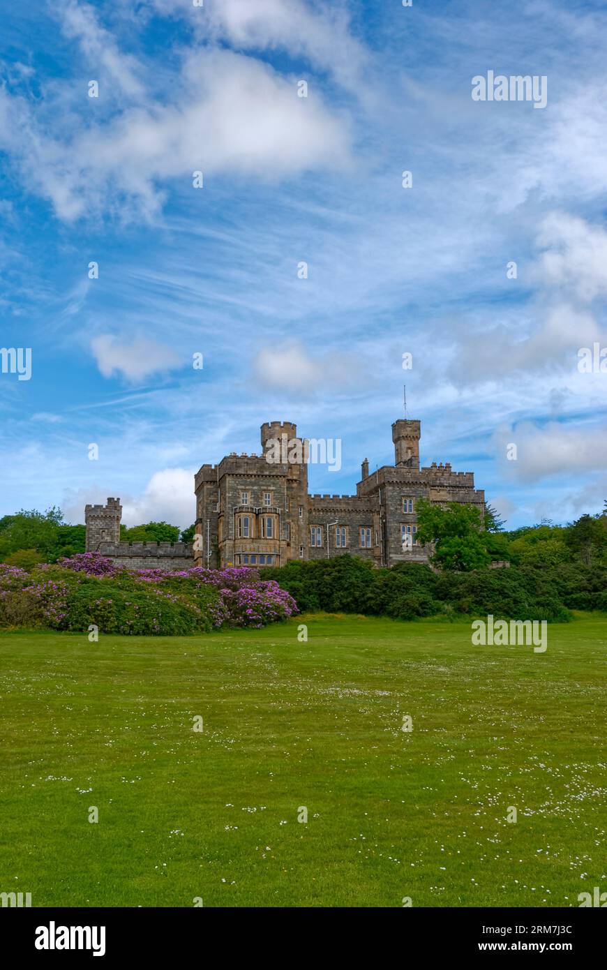 Lews Castle sul pendio erboso sopra Stornoway Town e parzialmente nascosto da alberi di rododendro fioriti sotto un cielo blu con nuvole sparse. Foto Stock