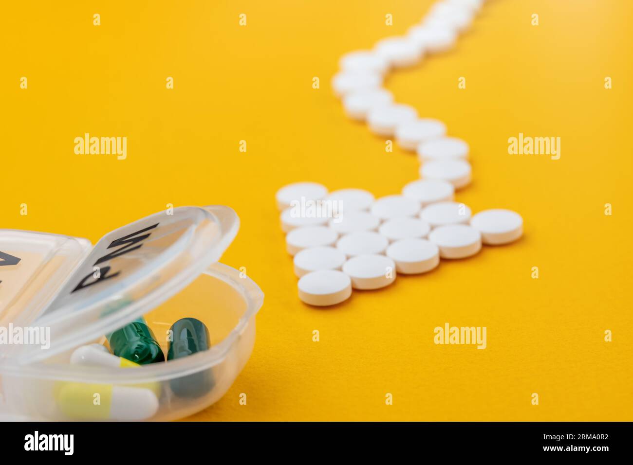 Pillole mediche bianche a forma di freccia puntano all'organizzatore quotidiano di plastica (per una settimana di farmaci) e fluiscono verso di esso. Primo piano su sfondo giallo. Foto Stock