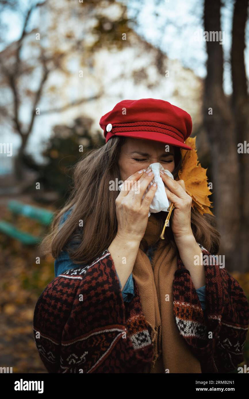 Ciao autunno. donna moderna di mezza età con cappello rosso con foglie autunnali, sciarpa e tovagliolo che soffiano il naso in città. Foto Stock