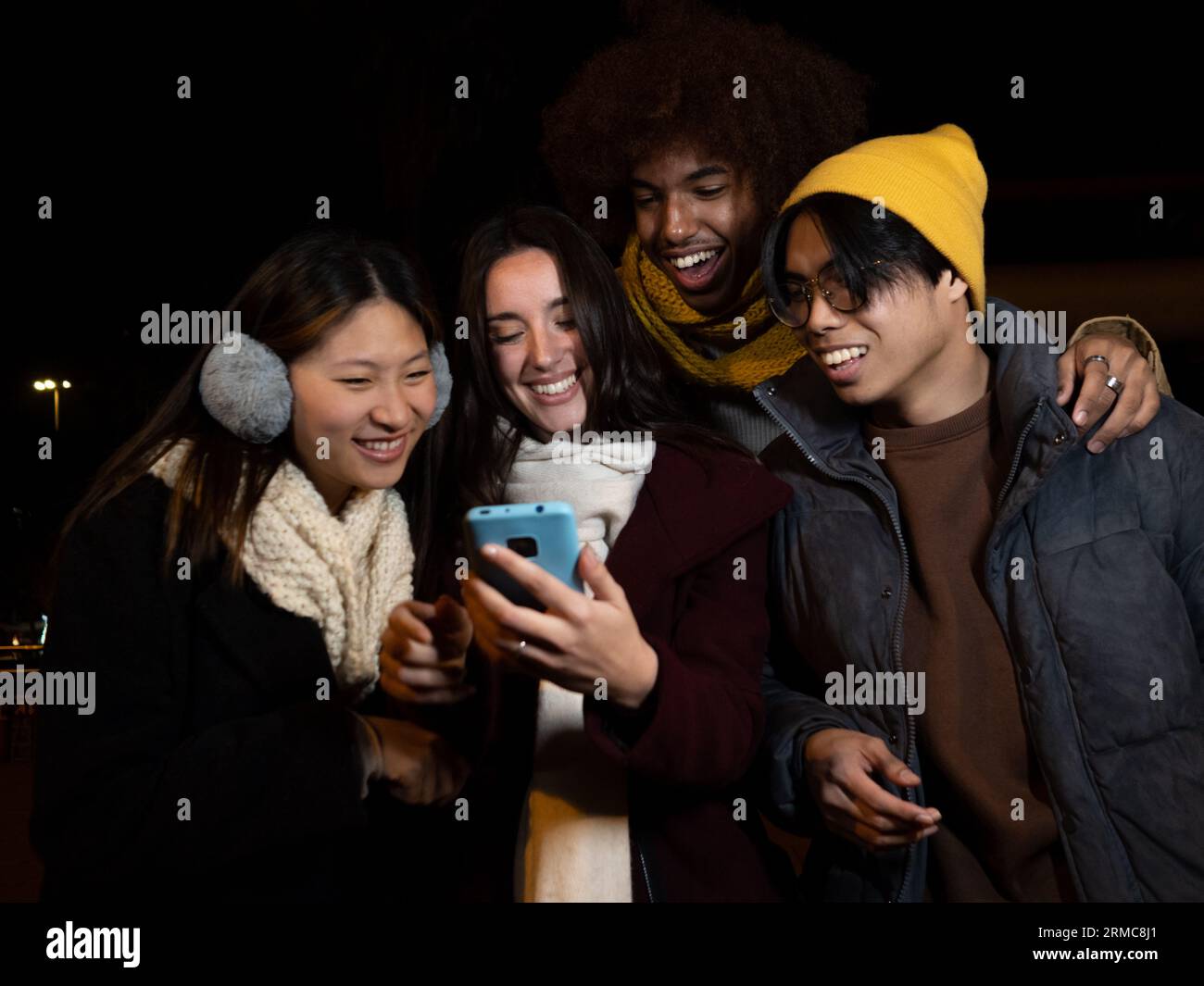 Gruppo di giovani diversi che si divertono con gli abiti invernali di notte in una città mentre naviga sul cellulare Foto Stock