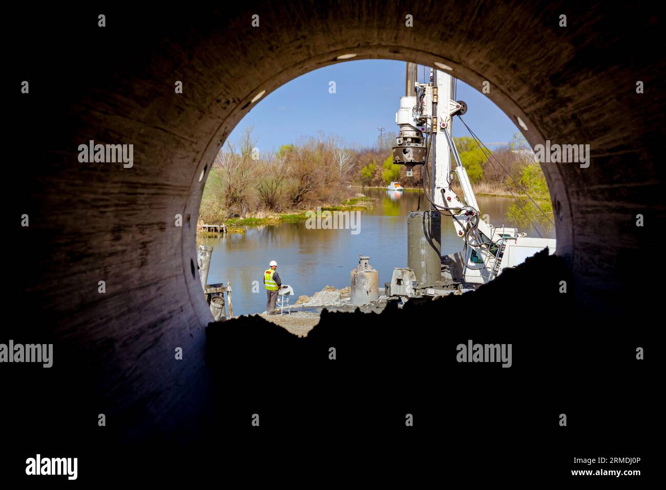 Vista in prospettiva del tubo a conca, da dietro sul lavoratore con giubbotto di sicurezza e casco bianco che gestisce la macchina di perforazione a torre, grandi attrezzature da perforare Foto Stock