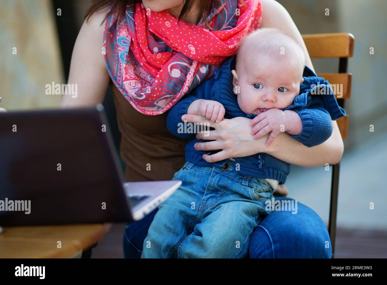Piccolo bambino di 4 mesi tra le braccia di sua madre mentre lavora al suo portatile Foto Stock
