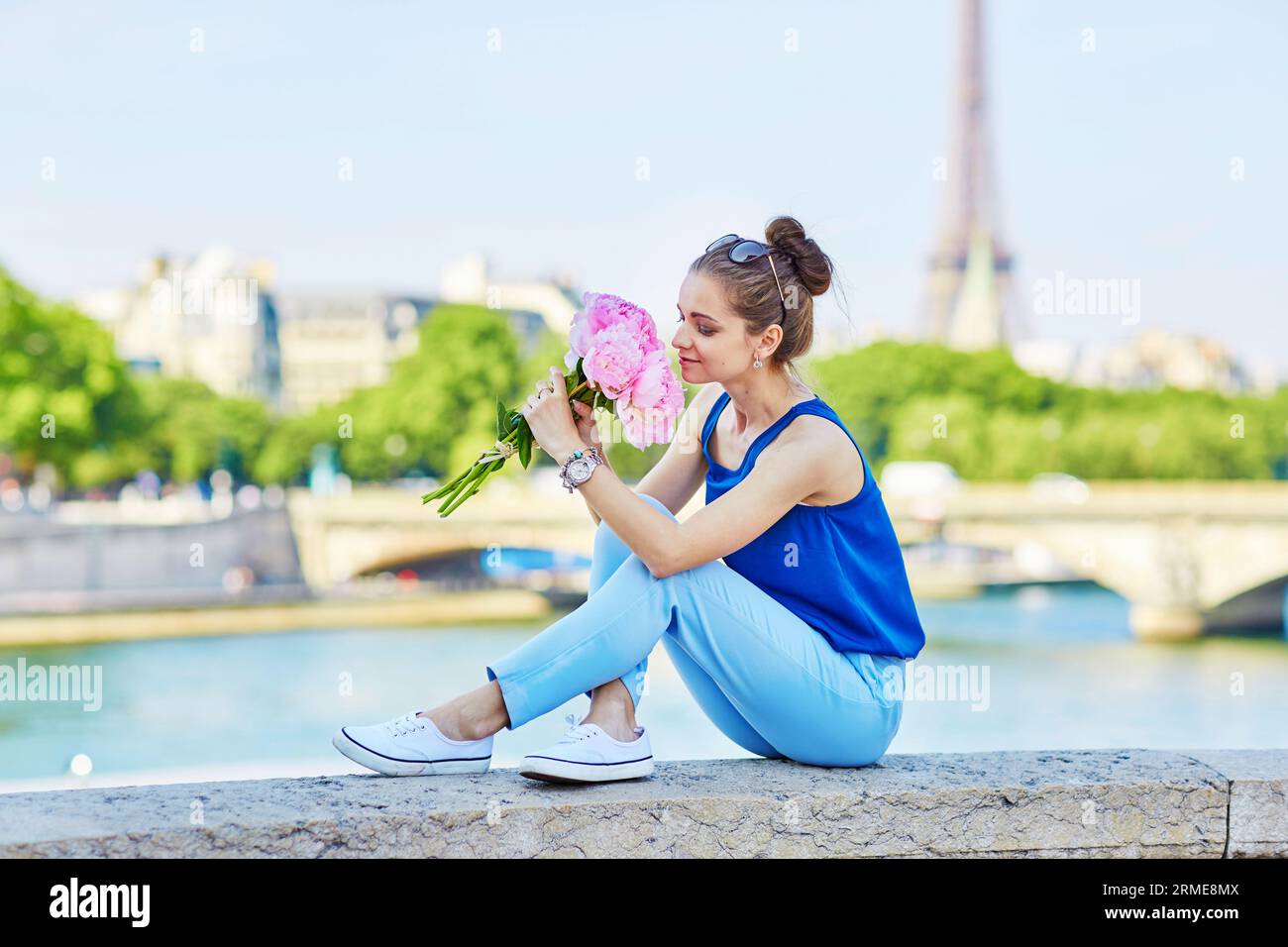 Bellissima giovane donna parigina in camicetta blu con un mucchio di peonie rosa sul ponte Alexandre III a Parigi, la Torre Eiffel è sullo sfondo Foto Stock