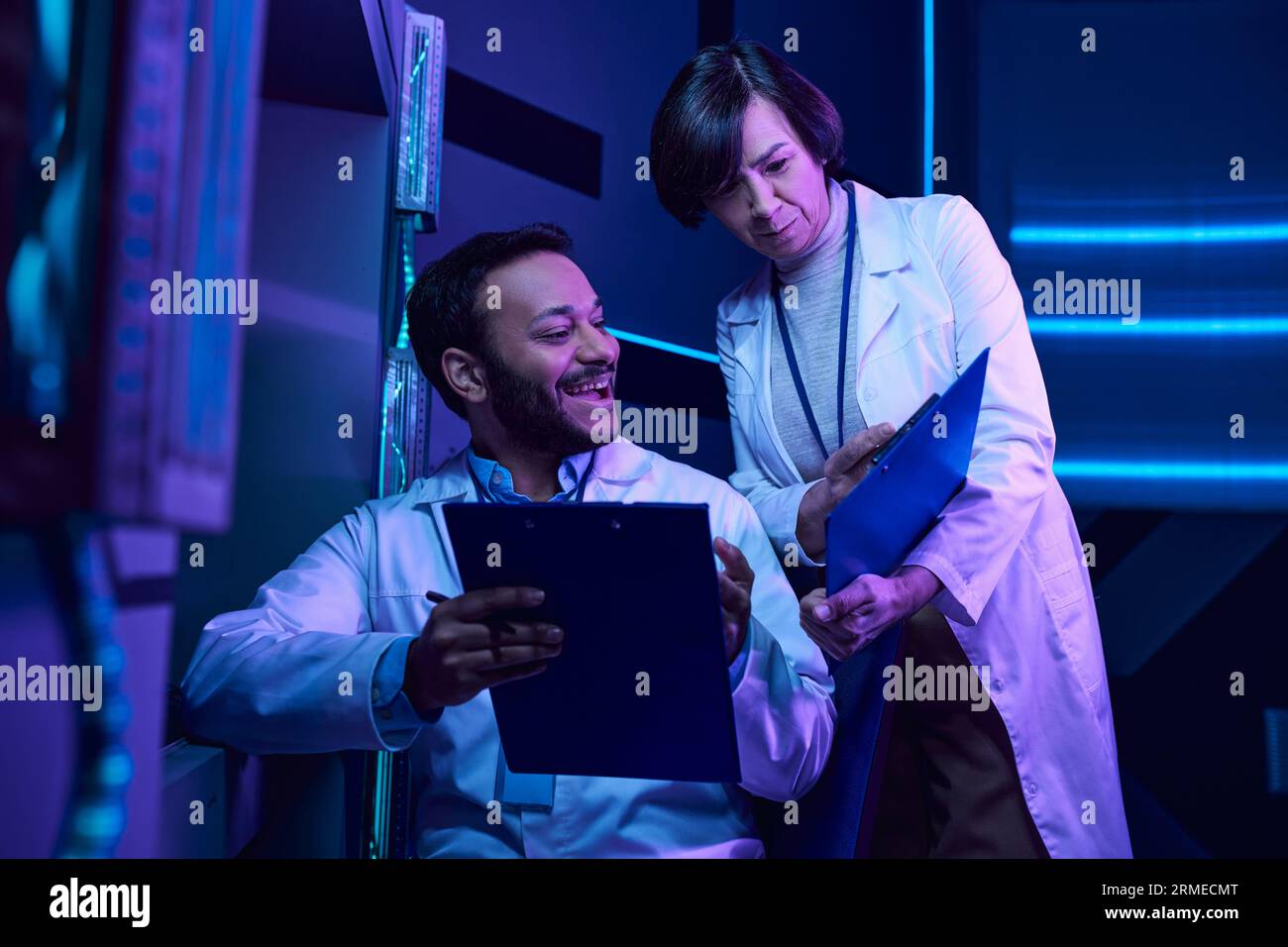 Scienziati indù maschi e adulti femmine analizzano i dati e condividono sorrisi nel Neon-Lit Science Center Foto Stock