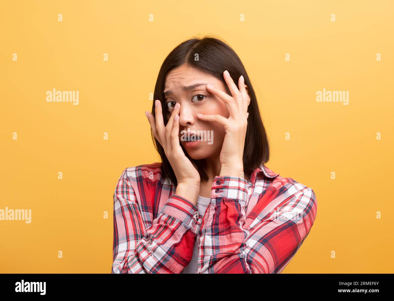 Una giovane studentessa cinese spaventata con un abito casual che le copre il viso con le mani e con uno sfondo giallo spaventato Foto Stock