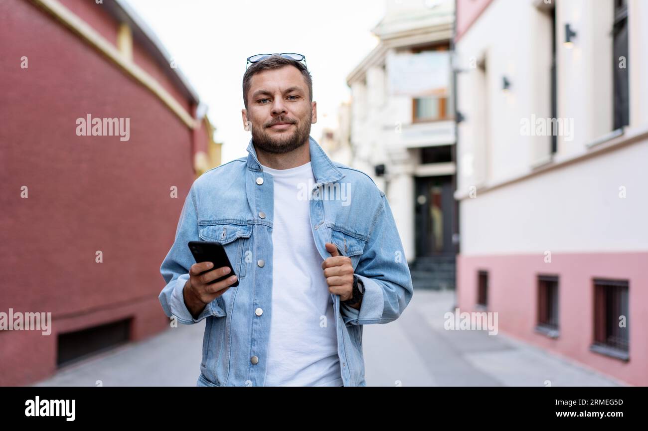 Giovane uomo adulto che indossa una giacca in denim di tendenza in piedi su un telefono in mano e sorride. Foto Stock