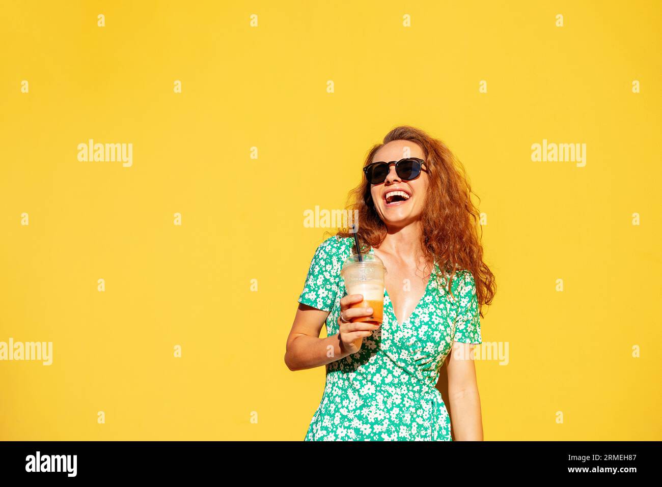 Allegra giovane donna dai capelli ricci con occhiali da sole che tiene in mano un bicchiere di succo e ride. Foto Stock