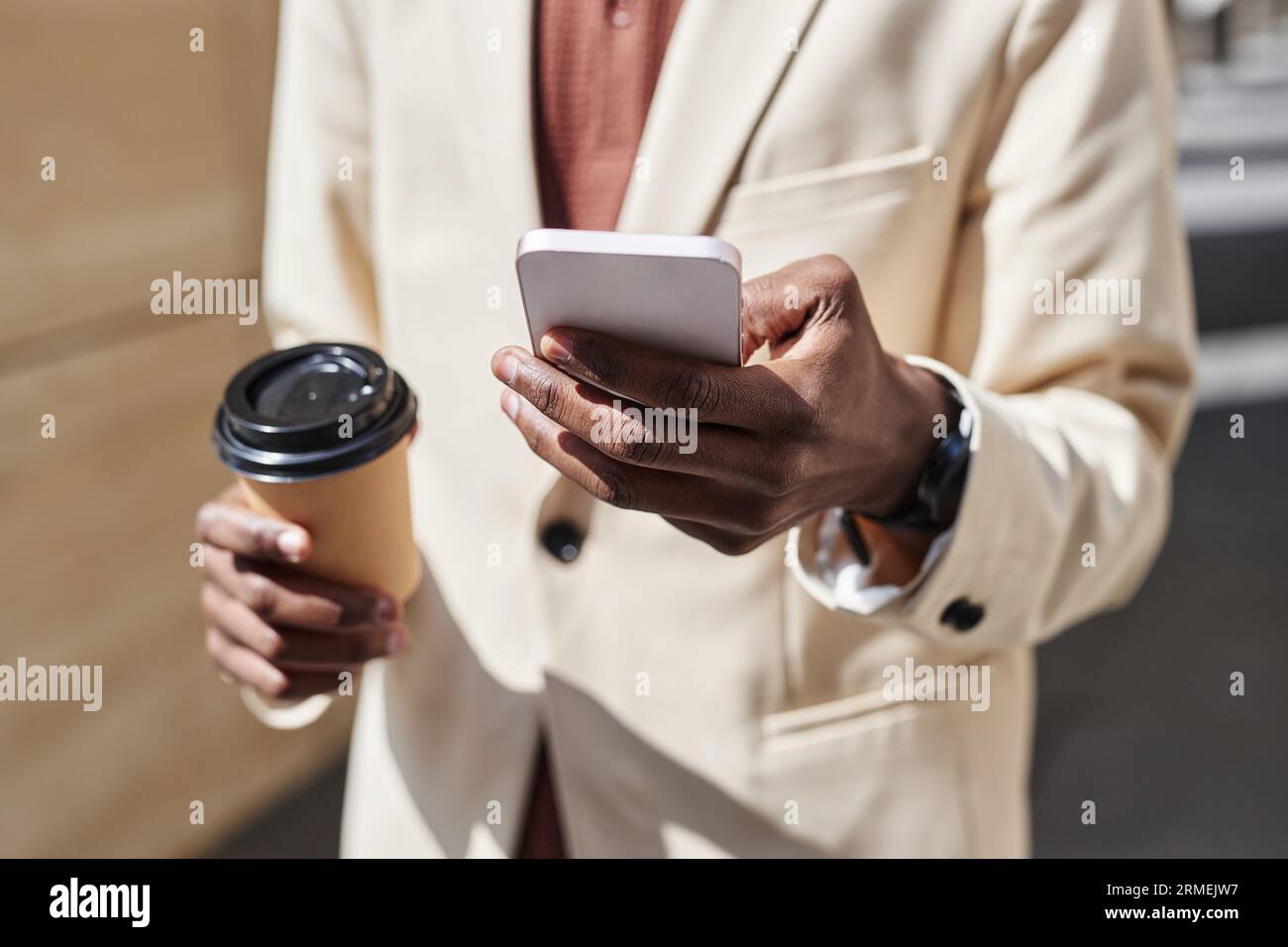 Concentrati sulla mano di un uomo d'affari con un abbigliamento in formalwear bianco che invia messaggi sullo smartphone e sorseggia un caffè davanti alla fotocamera nelle giornate di sole Foto Stock