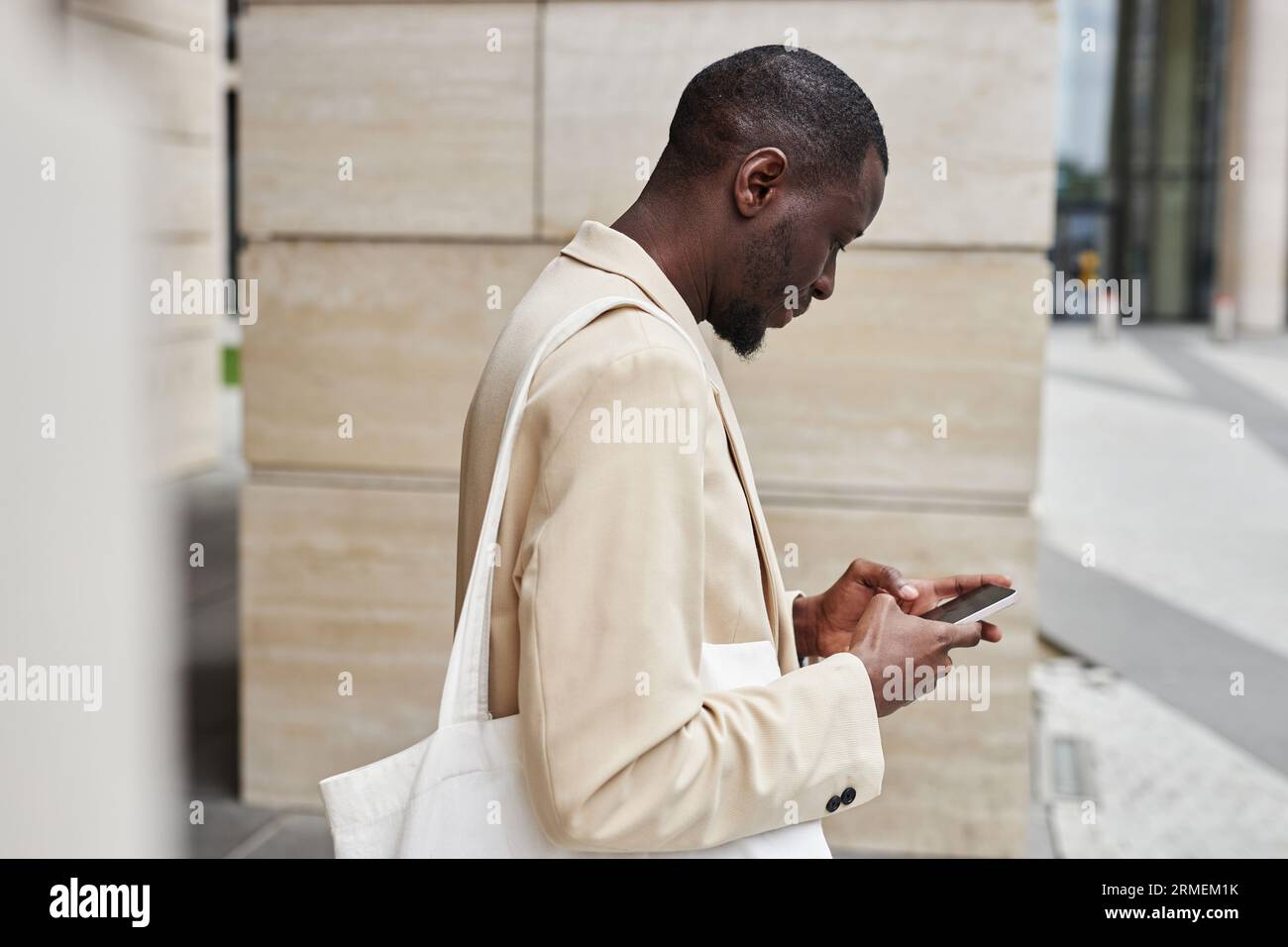 Direttore maschile afroamericano in formalwear bianco che invia messaggi sullo smartphone mentre si trova di fronte alla fotocamera di un edificio moderno in strada Foto Stock