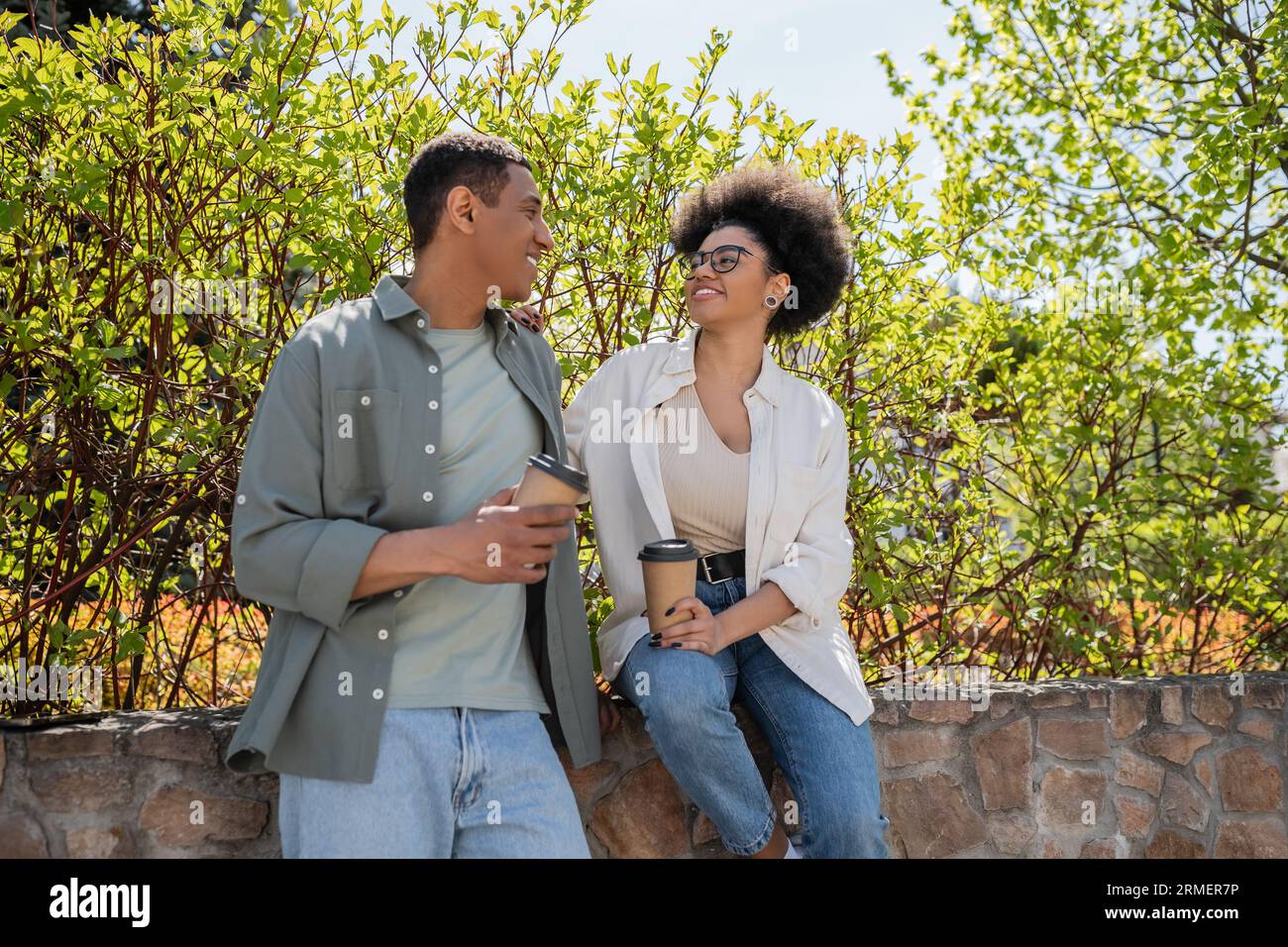 una coppia afro-americana positiva con un caffè per andare a parlare vicino agli alberi all'aperto in estate Foto Stock