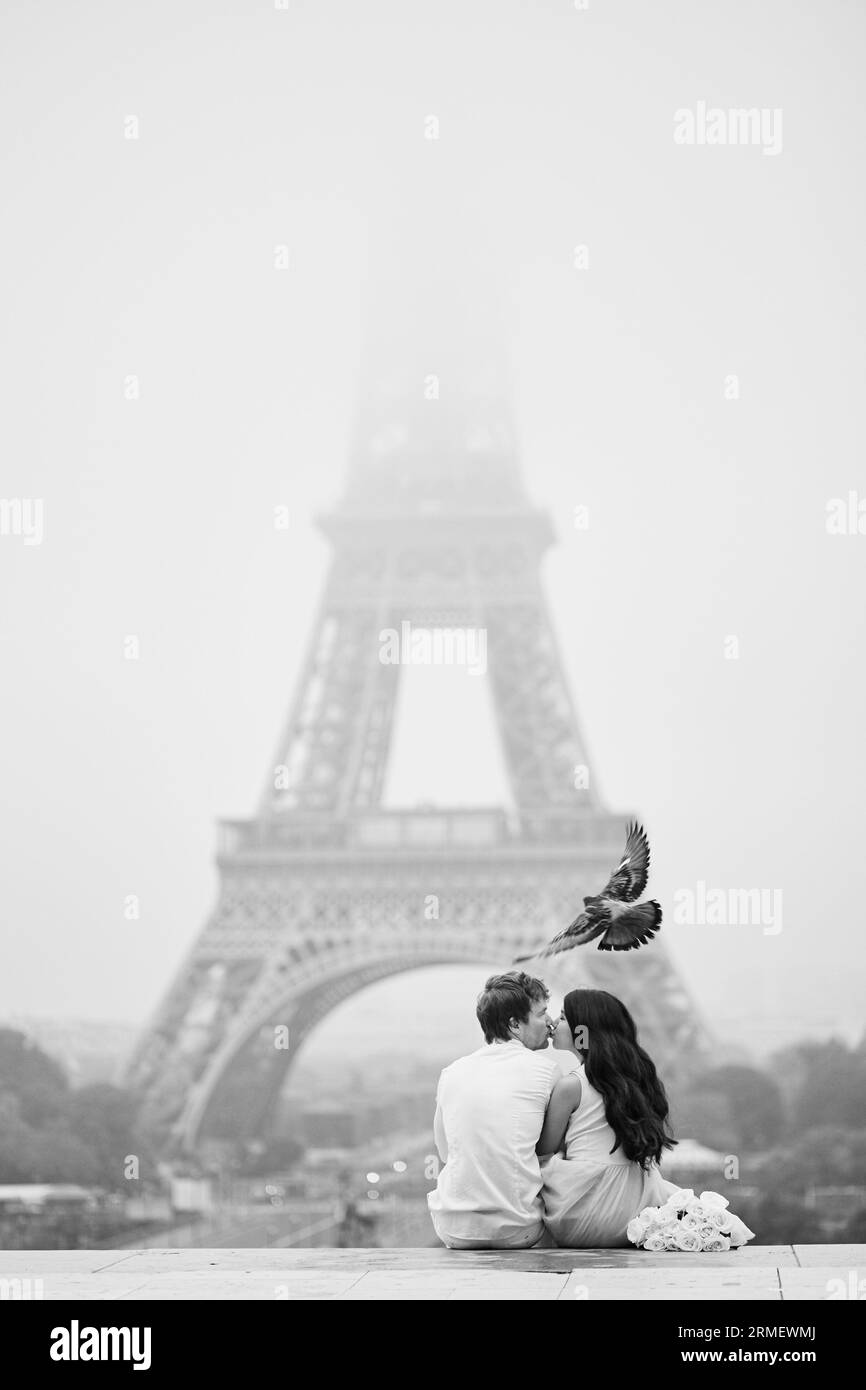 Bellissima coppia romantica innamorata di un mucchio di rose bianche sedute e baciate vicino alla Torre Eiffel a Parigi in una giornata piovosa e nebbiosa, uccello i. Foto Stock