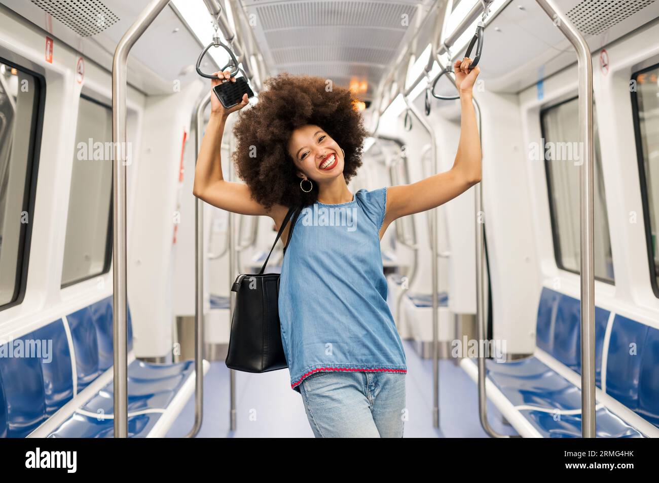 Allegra giovane passeggero di razza mista con capelli afro scuri e labbra rosse che sorridono ampiamente e guardano la macchina fotografica mentre si trova in piedi in un vagone ferroviario vuoto W. Foto Stock