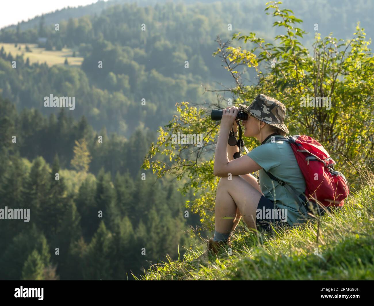 Una turista seduto su una collina, ammirando la vista delle montagne attraverso il binocolo Foto Stock