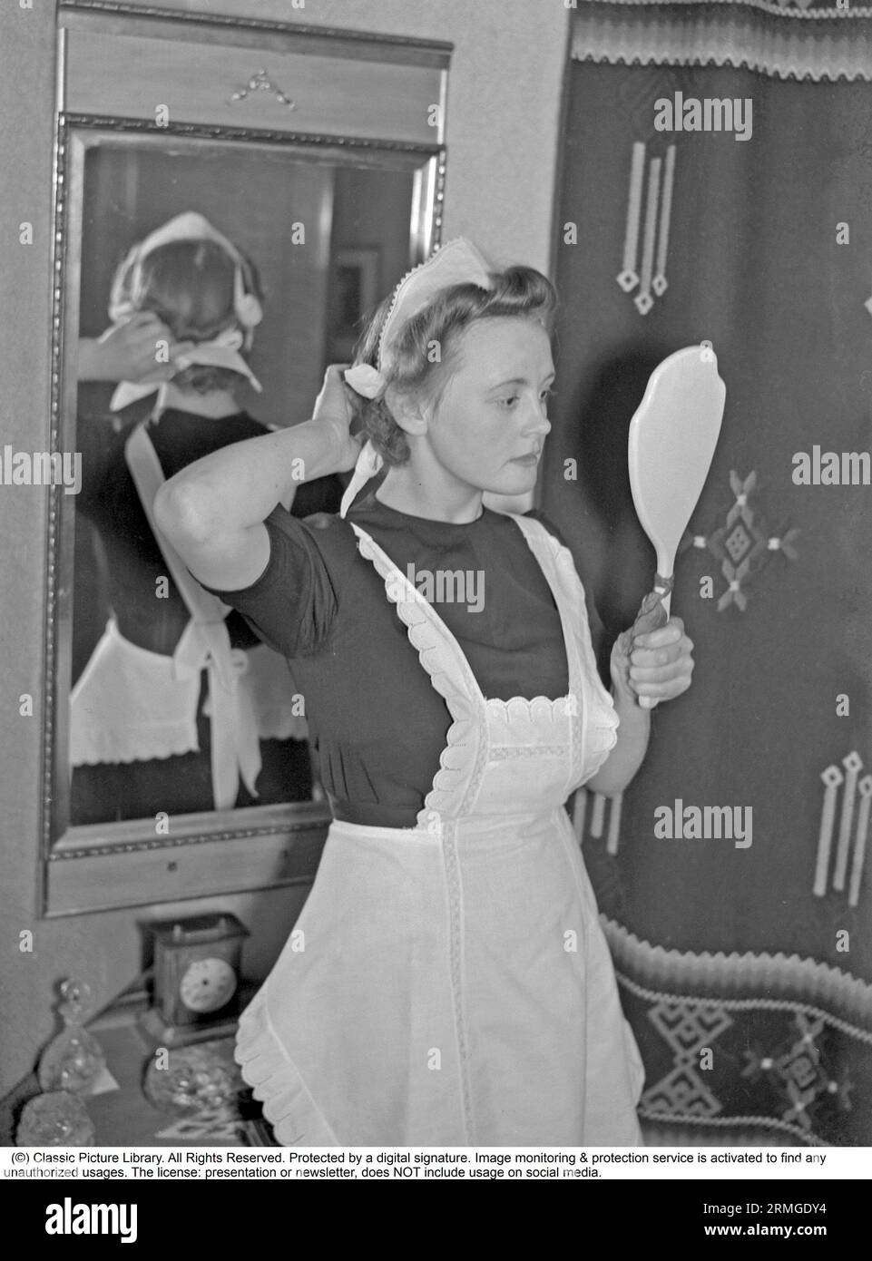 Negli anni '1940 Una giovane donna che lavora come cameriera si guarda nello specchio e mette i capelli in ordine e in ordine. Lei è vestita in modo ordinato con un grembiule bianco. Svezia 1940. Kristoffersson rif. 56-6 Foto Stock