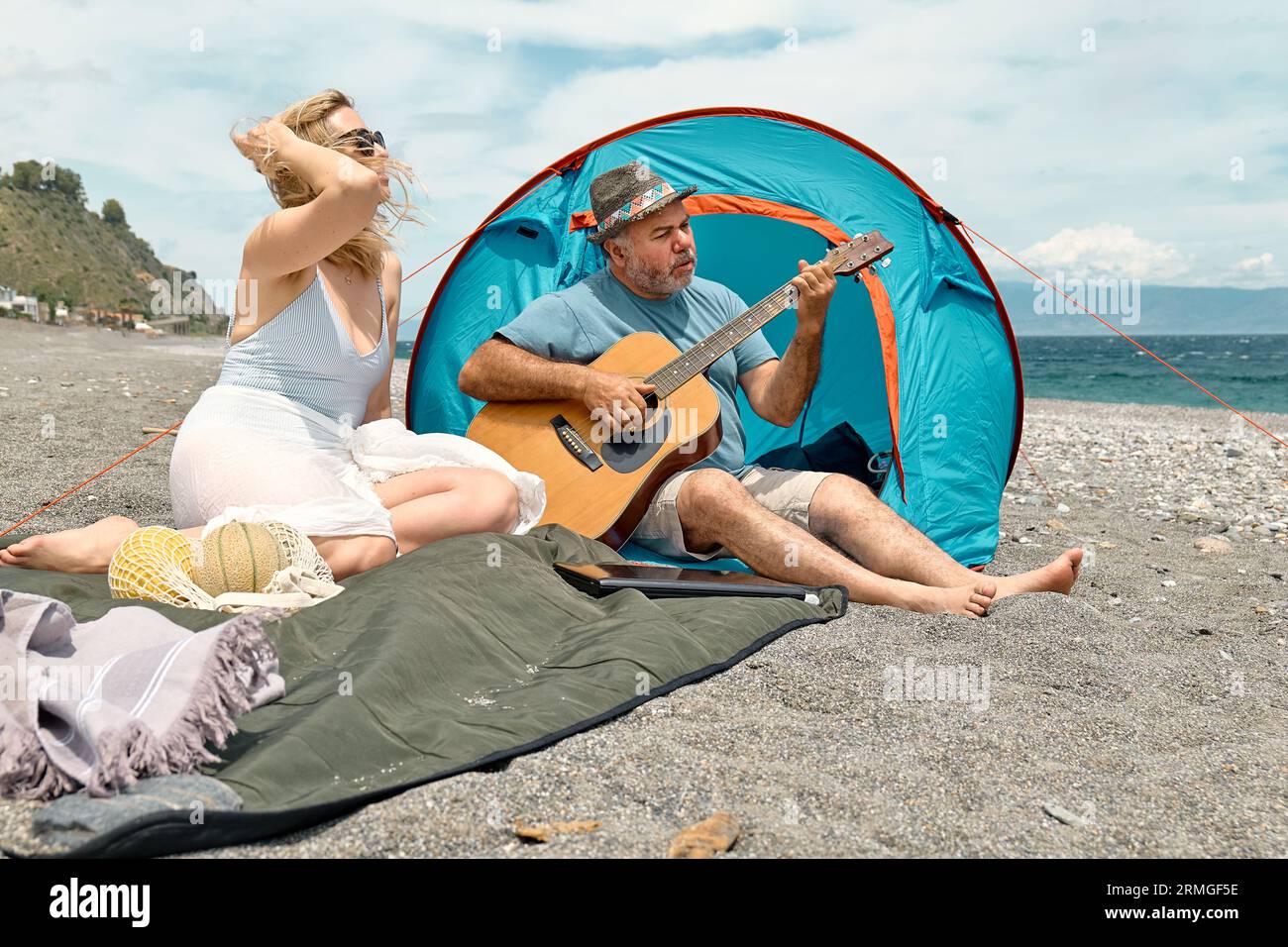 Coppia di mezza età che si diverte, canta e suona la chitarra in un picnic e campeggia sulla spiaggia durante le vacanze estive all'aperto. Viaggi, vacanze e amici Foto Stock