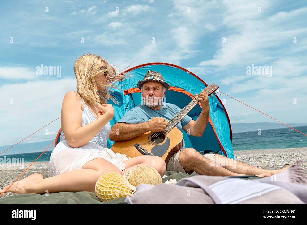 Coppia di mezza età che si diverte, canta e suona la chitarra in un picnic e campeggia sulla spiaggia durante le vacanze estive all'aperto. Viaggi, vacanze e amici Foto Stock