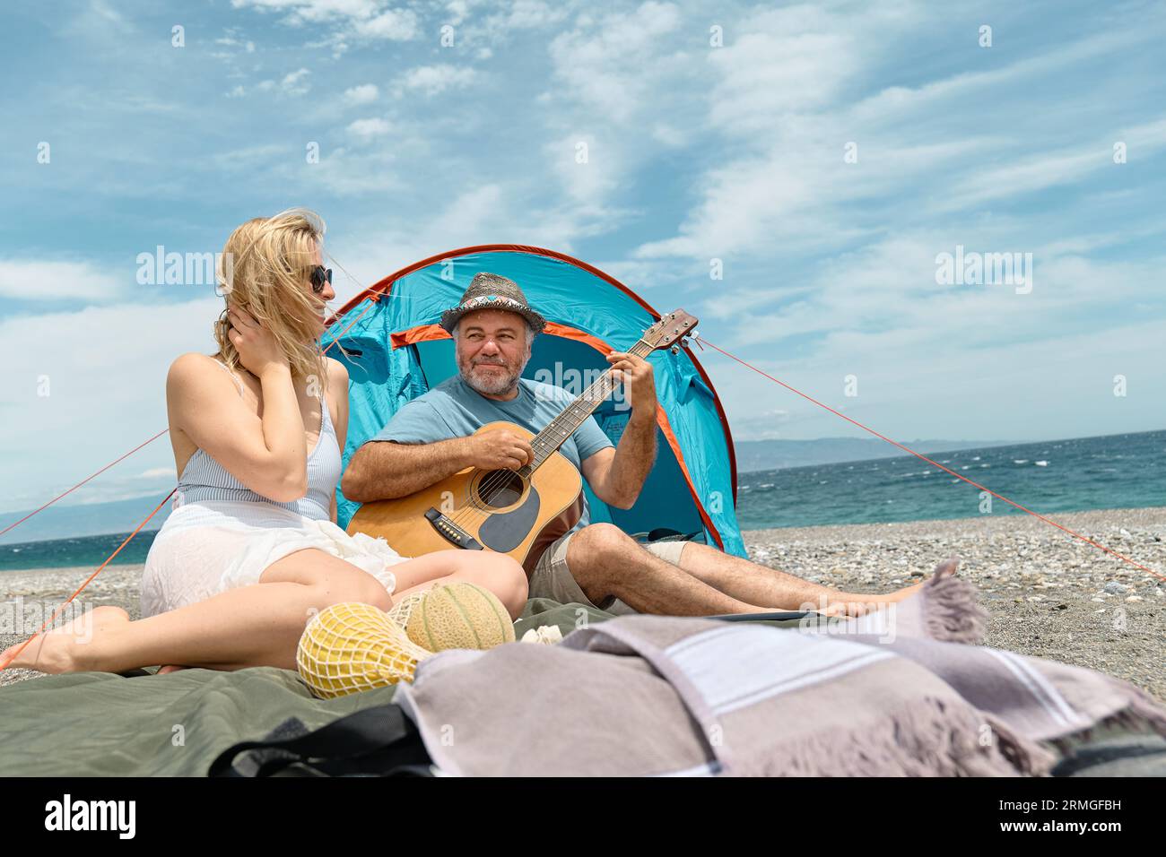 Coppia di mezza età che si diverte, canta e suona la chitarra in un picnic e campeggia sulla spiaggia durante le vacanze estive all'aperto. Viaggi, vacanze e amici Foto Stock