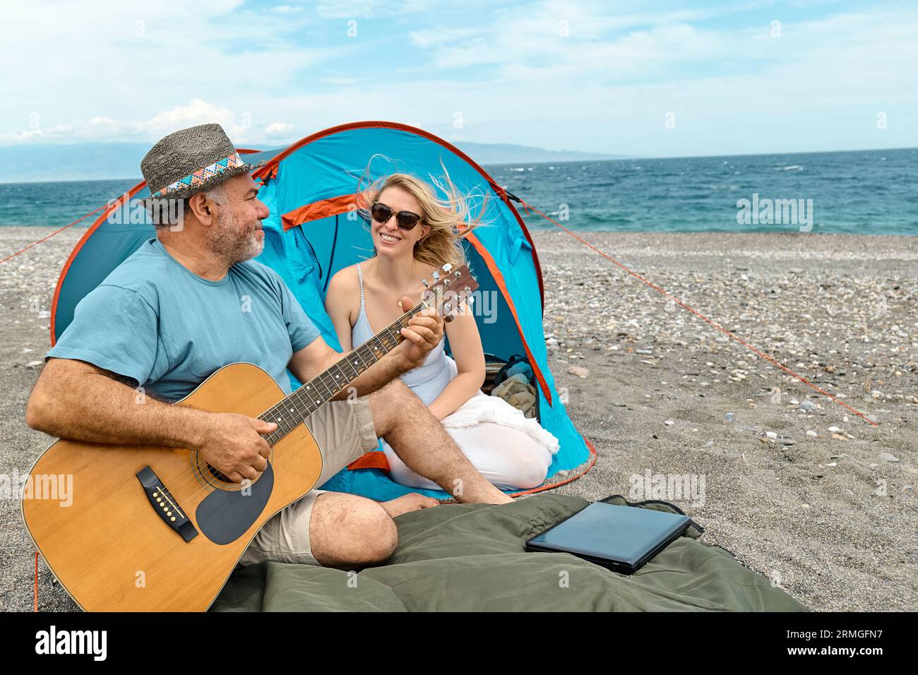 Coppia di mezza età che si diverte, canta e suona la chitarra in un picnic e campeggia sulla spiaggia durante le vacanze estive all'aperto. Viaggi, vacanze e amici Foto Stock