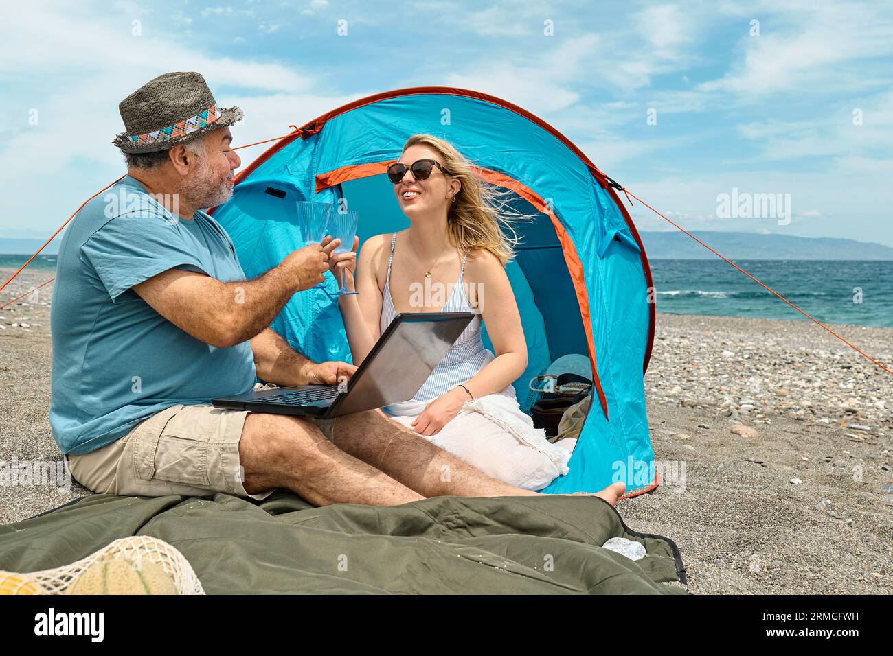 Coppia che si diverte, beve vino e parla con gli amici in videochiamata nel laptop in un picnic vicino a una tenda sul mare. Vacanze estive sulla spiaggia di Outd Foto Stock