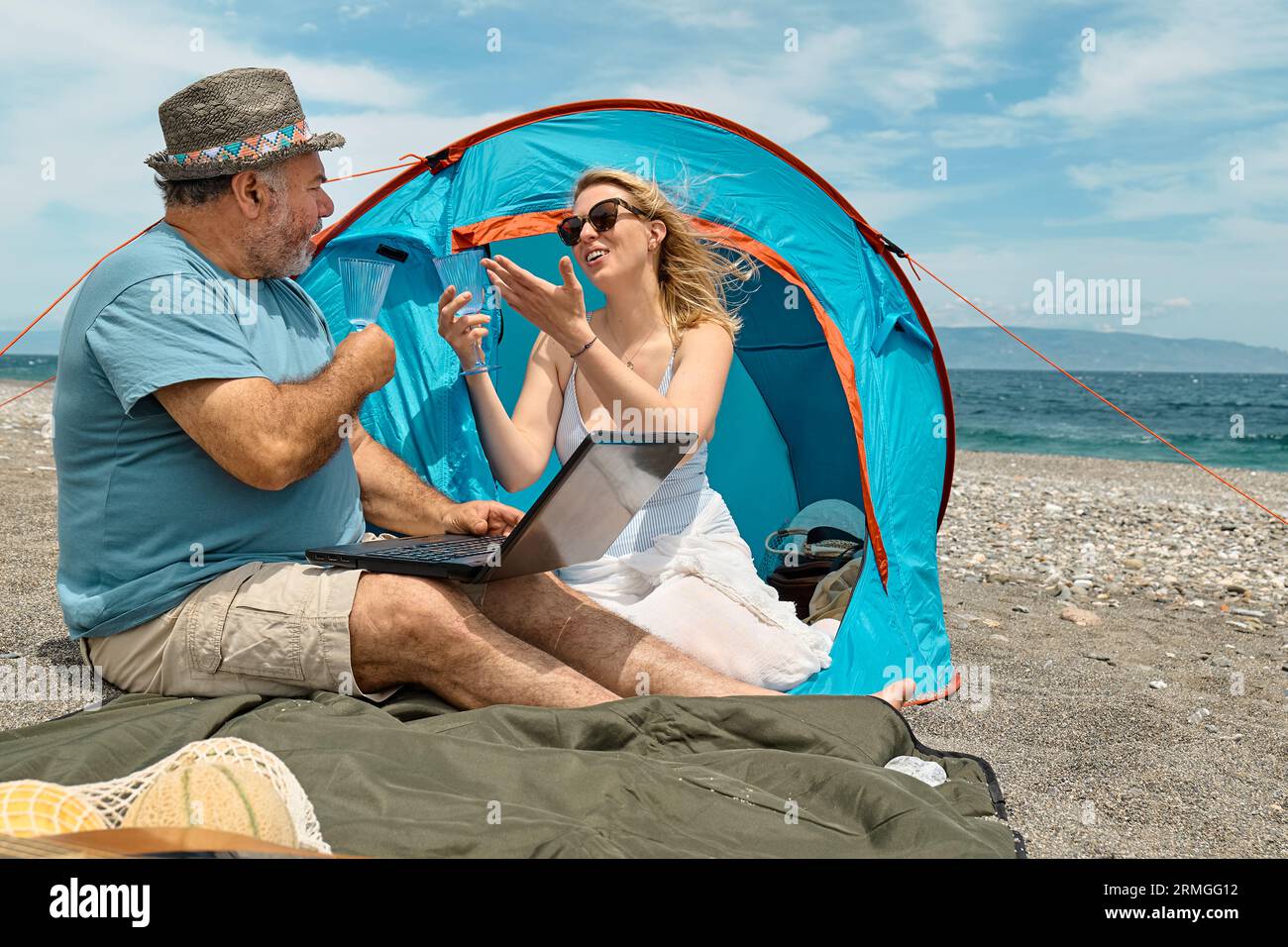 Coppia che si diverte, beve vino e parla con gli amici in videochiamata nel laptop in un picnic vicino a una tenda sul mare. Vacanze estive sulla spiaggia di Outd Foto Stock