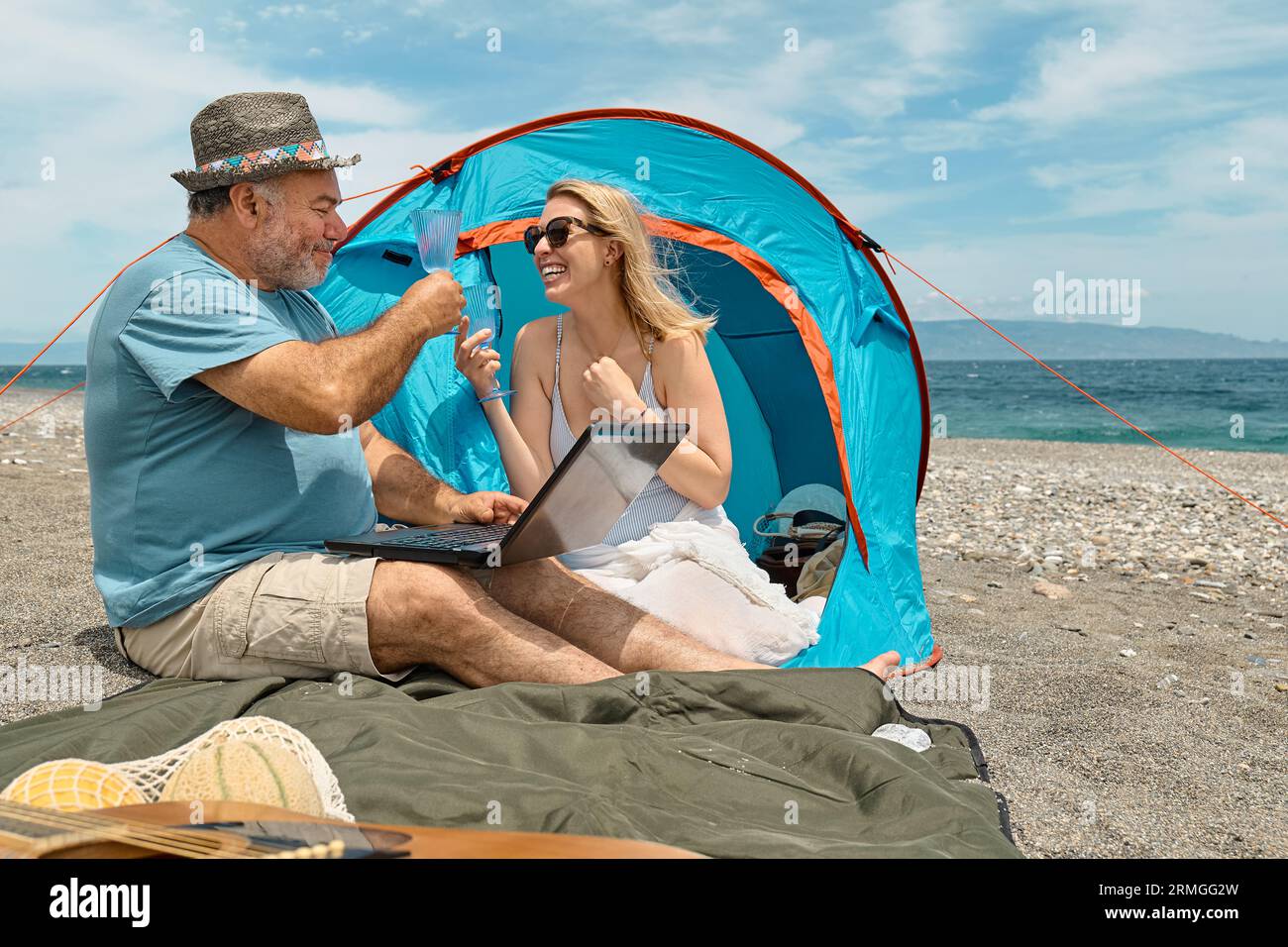 Coppia che si diverte, beve vino e parla con gli amici in videochiamata nel laptop in un picnic vicino a una tenda sul mare. Vacanze estive sulla spiaggia di Outd Foto Stock