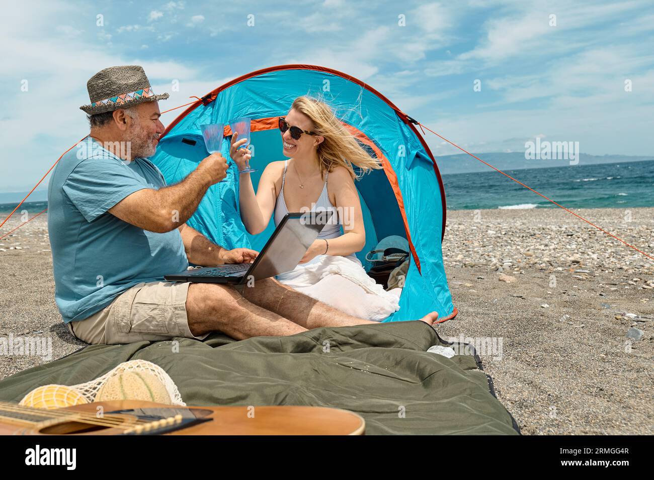 Coppia che si diverte, beve vino e parla con gli amici in videochiamata nel laptop in un picnic vicino a una tenda sul mare. Vacanze estive sulla spiaggia di Outd Foto Stock