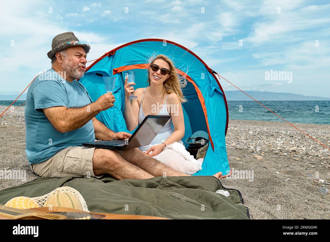 Coppia che si diverte, beve vino e parla con gli amici in videochiamata nel laptop in un picnic vicino a una tenda sul mare. Vacanze estive sulla spiaggia di Outd Foto Stock