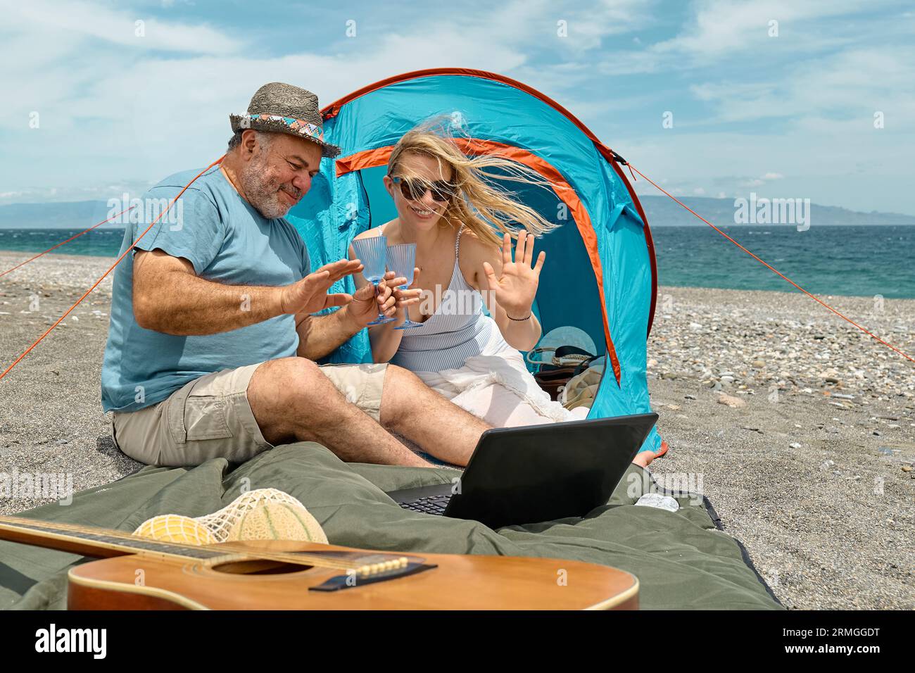 Coppia che si diverte, beve vino e parla con gli amici in videochiamata nel laptop in un picnic vicino a una tenda sul mare. Vacanze estive sulla spiaggia di Outd Foto Stock