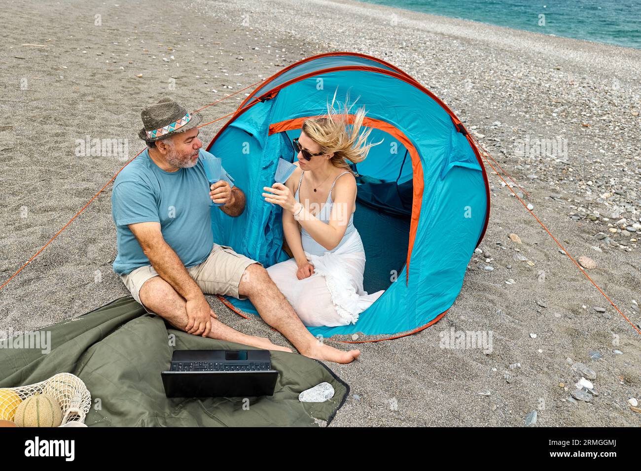 Coppia che si diverte, beve vino e parla con gli amici in videochiamata nel laptop in un picnic vicino a una tenda sul mare. Vacanze estive sulla spiaggia di Outd Foto Stock