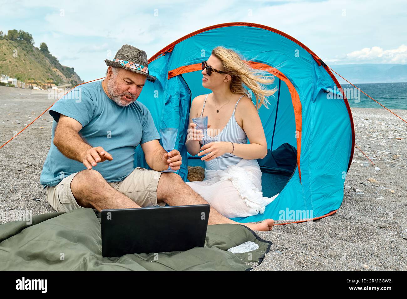 Coppia che si diverte, beve vino e parla con gli amici in videochiamata nel laptop in un picnic vicino a una tenda sul mare. Vacanze estive sulla spiaggia di Outd Foto Stock